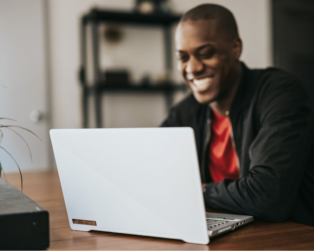Um homem com uma expressão alegre está sentado em uma mesa, concentrado em seu trabalho em um laptop.